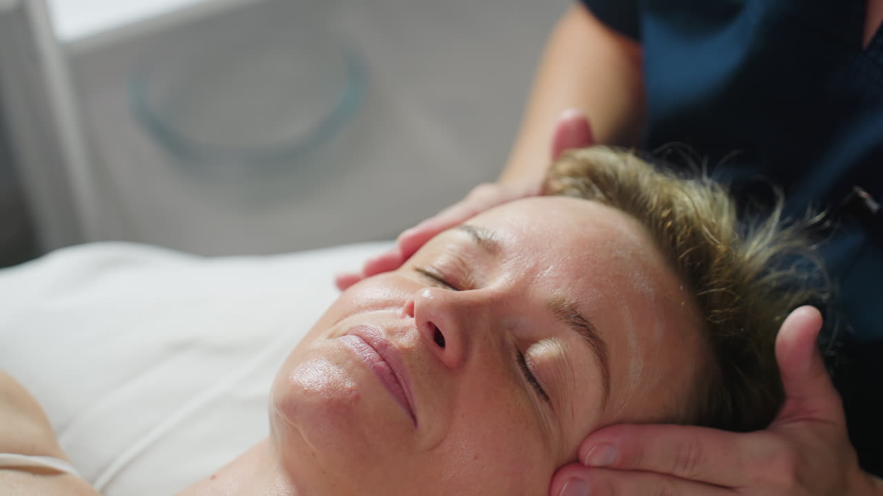 Recipient lies relaxed with eyes closed while skin therapist gently massages product across cheeks and forehead, creating glowing hydrated complexion in tranquil spa treatment setting