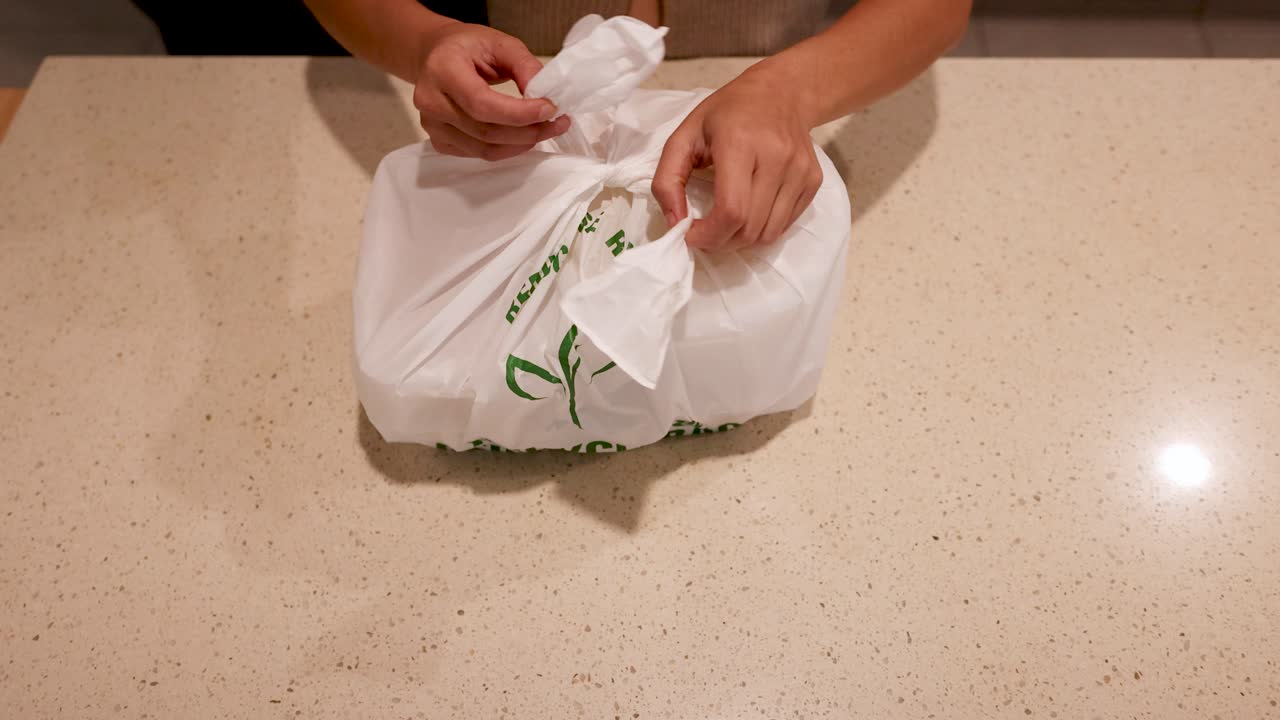 Hands open a recyclable plastic bag on a countertop, revealing food containers. Warm lighting enhances the eco-friendly theme