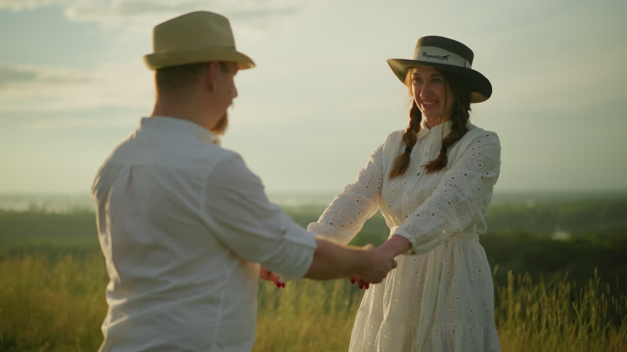 A couple stands in a sunlit grassland, holding hands and gazing lovingly at each other. Both are dressed in white and wearing hats, their moment captured beautifully in the golden light of sunset