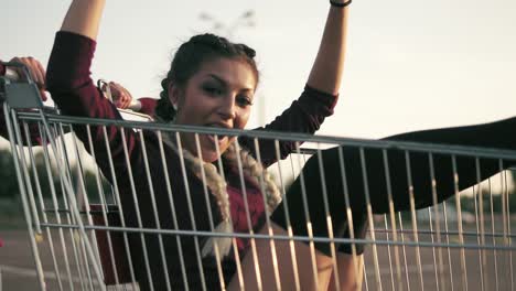 attractive smiling woman is sitting in the grocery cart with her arms raised up, while her friend is pushing her behind during