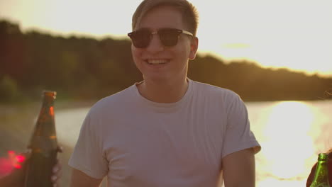 a brutal young man celebrates a birthday with the best friends. this is carefree open air party with beer on the beach near the lake coast at sunset.