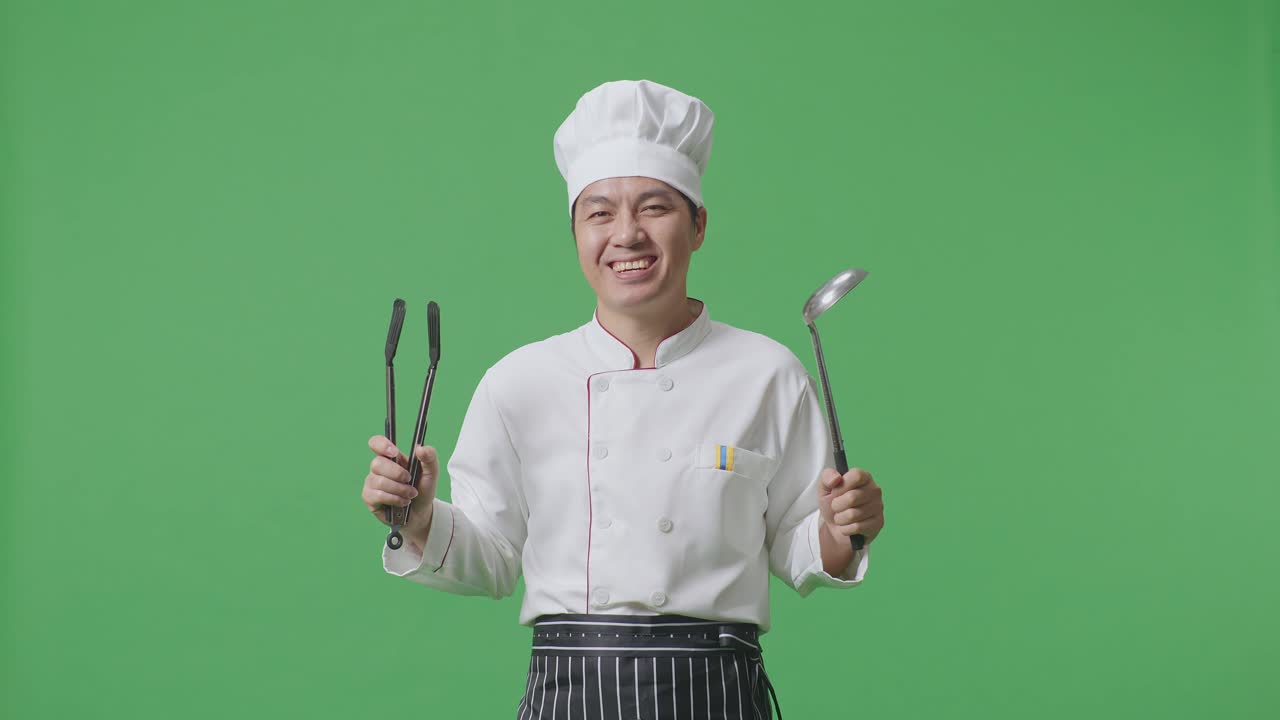 chef asiático sonriendo y mostrando pinzas y cuchara a la cámara mientras está de pie en el estudio de fondo de la pantalla verde