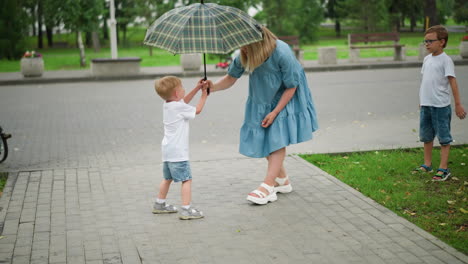 a mother holds an umbrella while her younger son joyfully tries to grab it, creating a playful moment, the older boy watches from behind