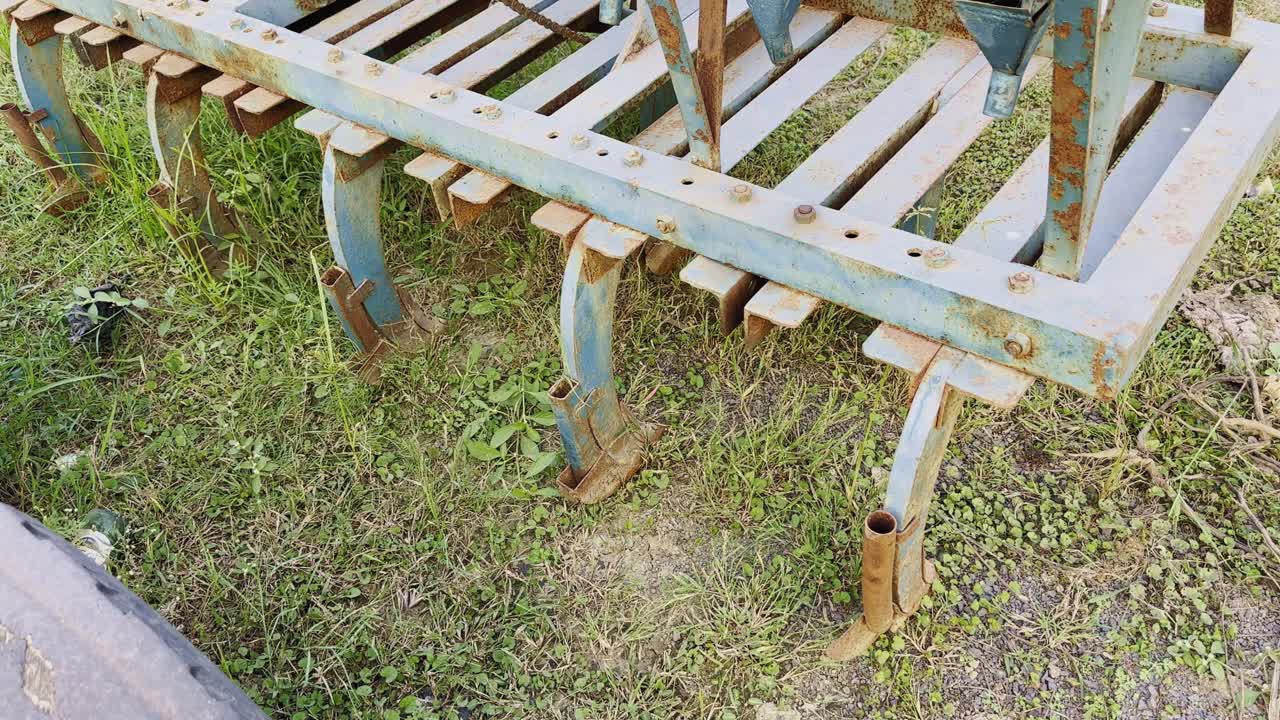 A smooth tracking shot showcasing an old rusty blue cultivator, its metal tines covered in dust and corrosion, resting on grass in an open farm field, symbolizing aging agricultural equipment