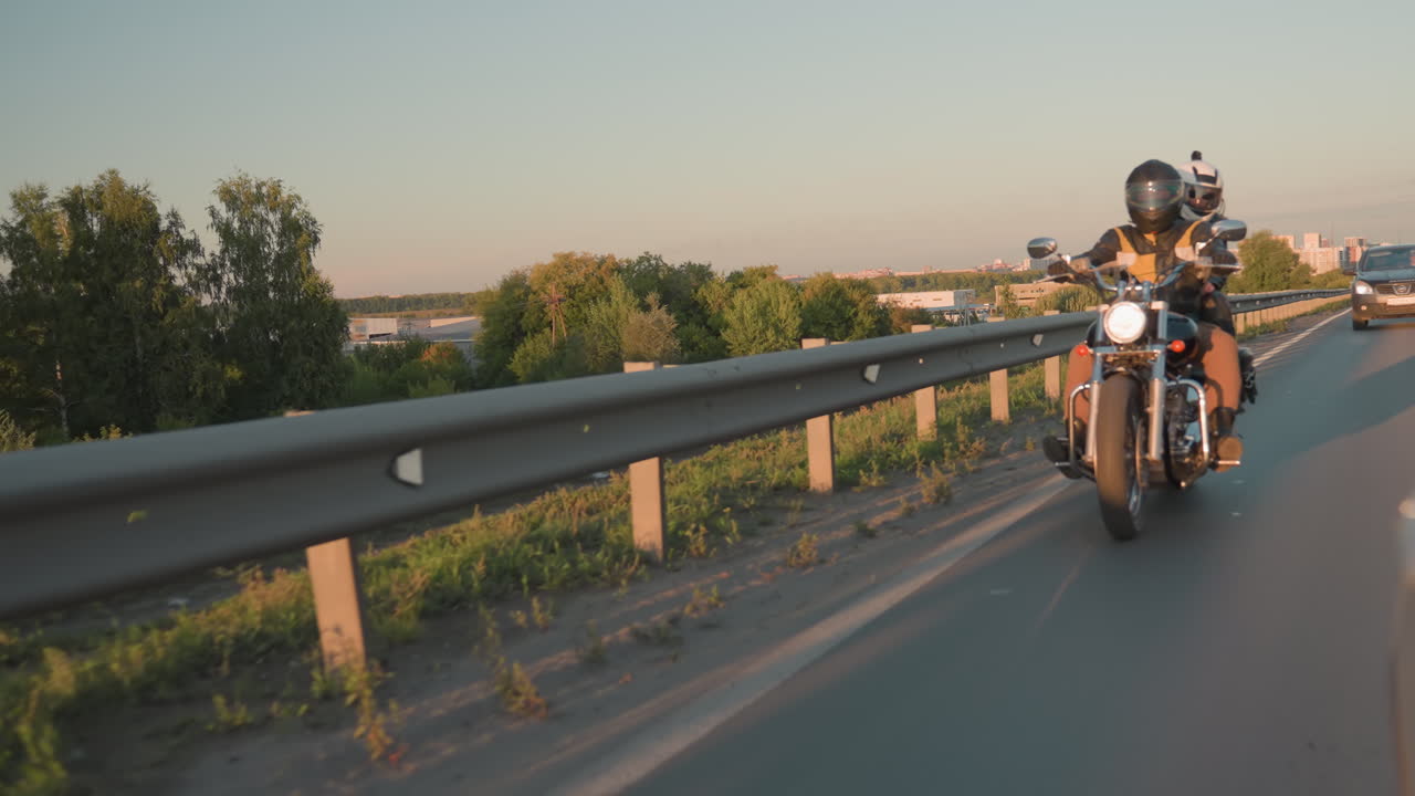 Folk som kjører motorsykkel nærme fortauet under «golden hour», med biler som følger bak, viser spenningen ved fart, vennskap, livsstil, eventyr og friheten på landeveien