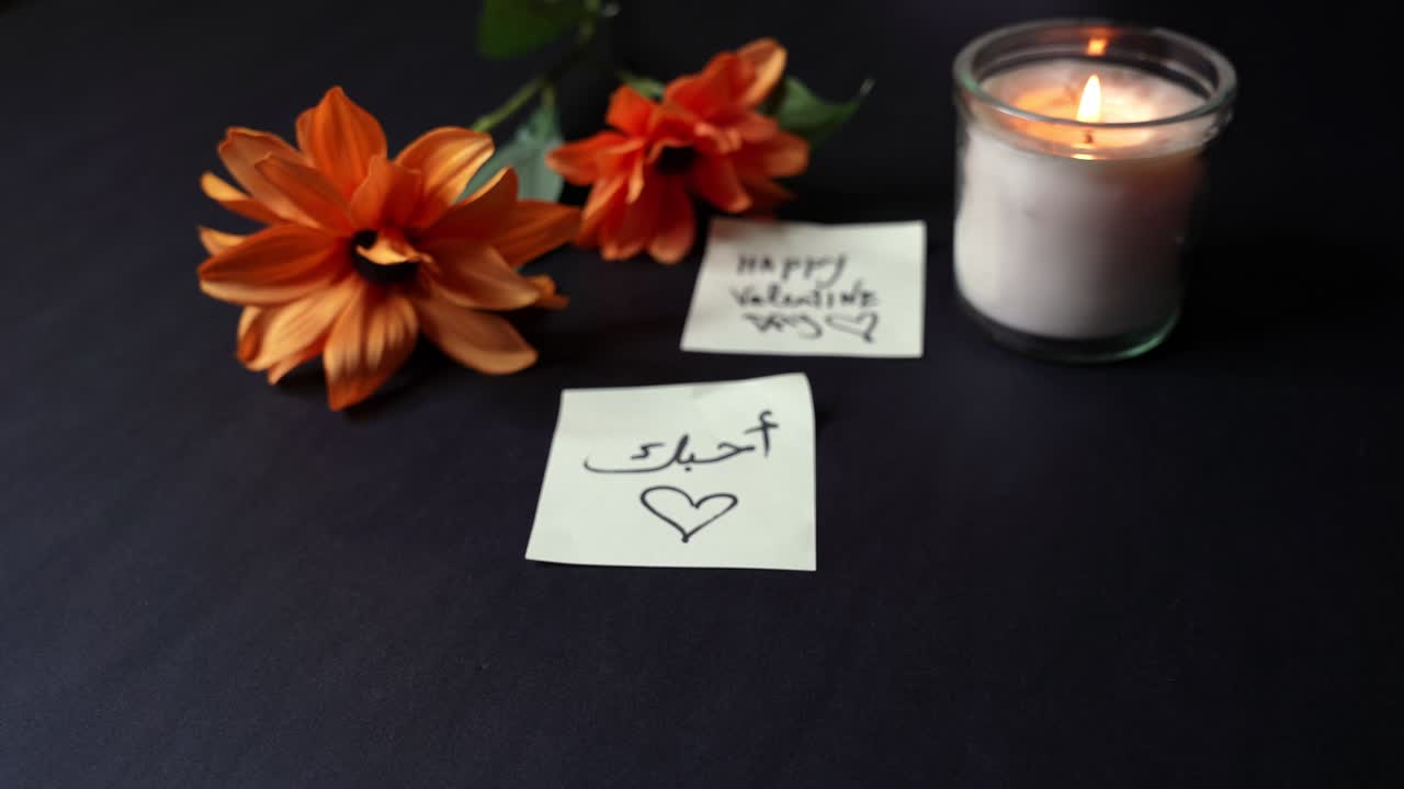 Nærbillede af en hånd, der holder en arabisk “Jeg elsker dig”-noteseddel nær et tændt glaslys og orange blomster på et mørkt bord, varmt stearinlys skaber romantisk Valentinsdagsstemning indendørs