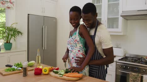 Lächelndes-Afroamerikanisches-Paar-Mit-Schürzen,-Das-Sich-In-Der-Küche-Umarmt-Und-Essen-Zubereitet