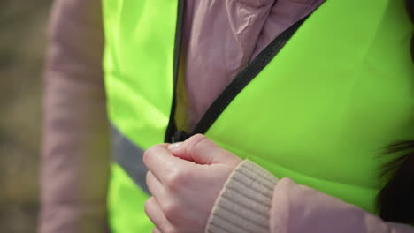 close-up of person wearing unzipped neon yellow reflective safety vest over pink padded jacket outdoors, showcasing bright high-visibility material and grey reflective stripes, in daylight
