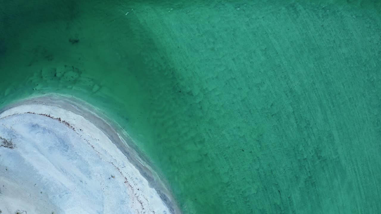 A sweeping arc of pale sand blends into luminous turquoise water where shifting tones and gentle contours create an abstract coastal pattern shaped by tide and shoreline flow