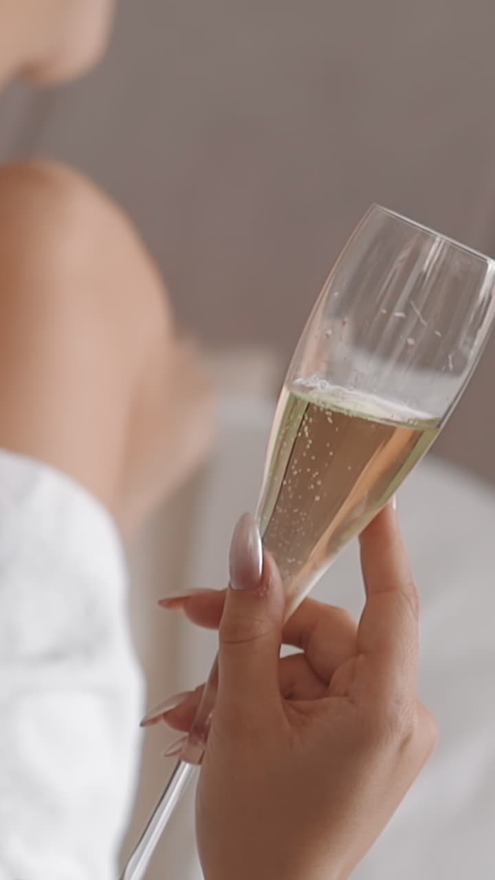 Lady wearing white bathrobe holds glass of champagne and swings legs on bed in hotel room close view. Holiday celebration at vip-resort. Luxury life
