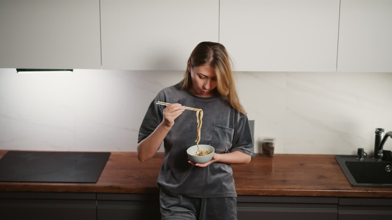 Young woman wearing gray casual outfit standing in modern kitchen holding bowl and eating pasta with chopsticks, looking down at meal with relaxed and focused expression