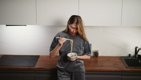 young woman wearing gray casual outfit standing in modern kitchen holding bowl and eating pasta with chopsticks, looking down at meal with relaxed and focused expression