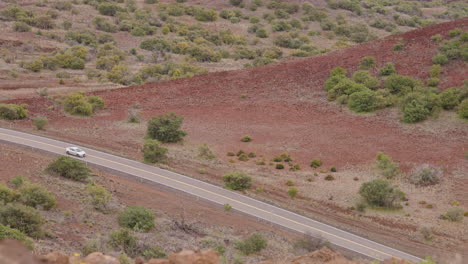 Slow-Motion-Car-On-Road-With-Red-Dirt-Vegetation-In-Valley-From-Mauna-Kea-Lookout,-Hawaii-4K