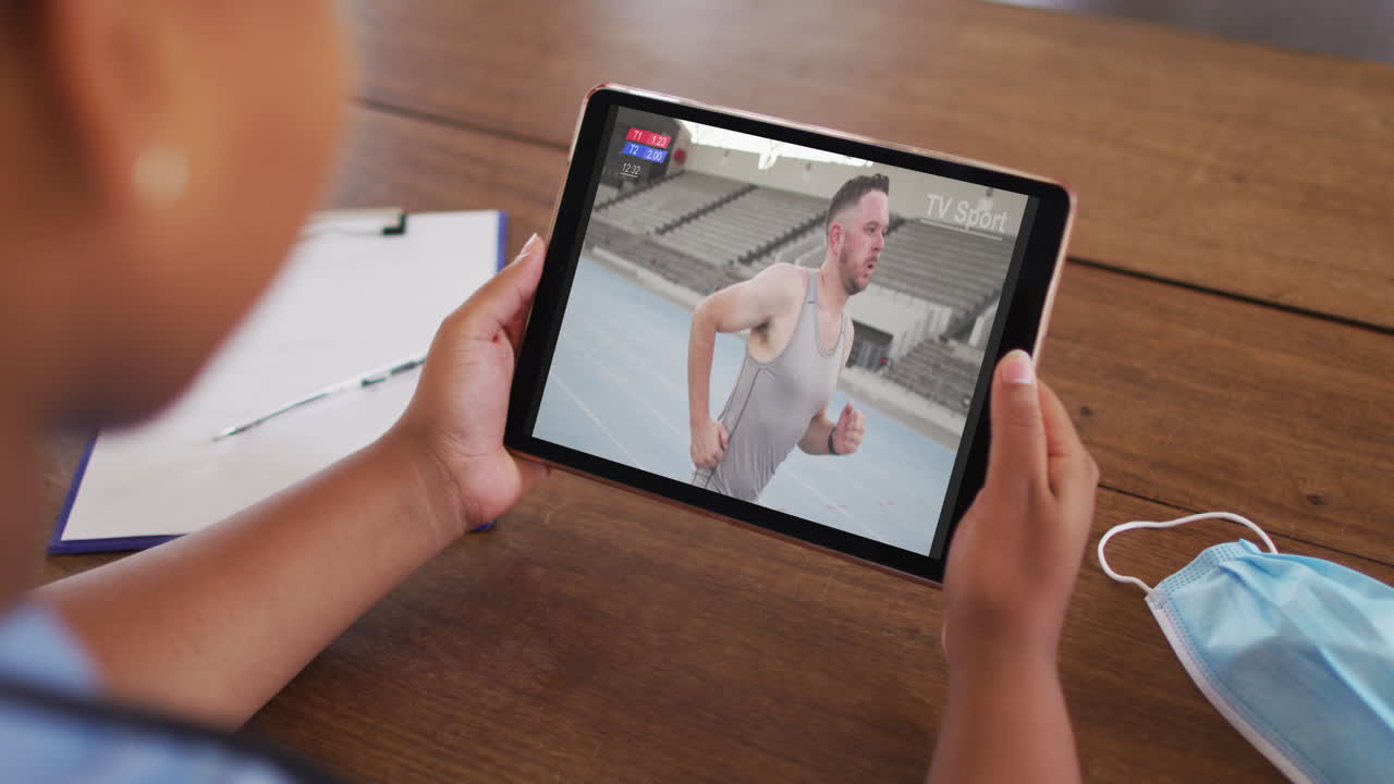 compuesto de una mujer sentada en una mesa con una máscara facial, viendo un evento atlético en la tableta