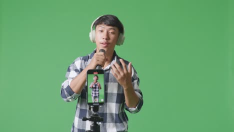móvil en vivo de un joven adolescente asiático con auriculares y sosteniendo un micrófono y cantando en el fondo de la pantalla verde