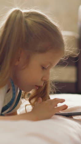 cu, tracking: little girl, beautiful, in blue sundress, with long pigtails, lies on floor and works on smartphone, child swipe, tap on screen, closeup