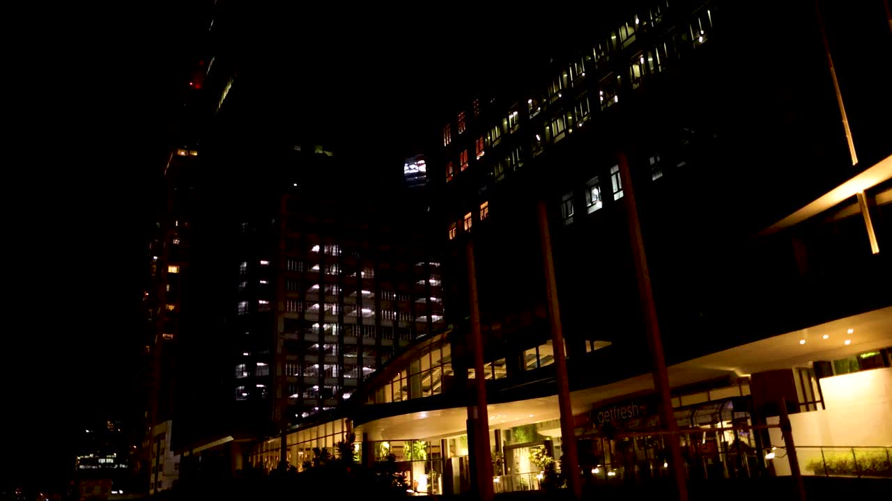 Camera pans upward revealing modern Bangkok skyscrapers and streetlights at night, low-light urban environment