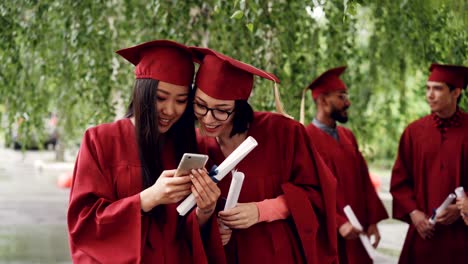 female graduates are using smartphone looking at screen talking and laughing standing outdoors holding diplomas, girls are wearing formal gowns and hats.