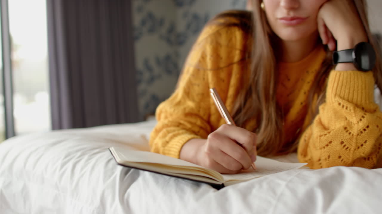 Writing in journal, woman in yellow sweater relaxing on bed at home