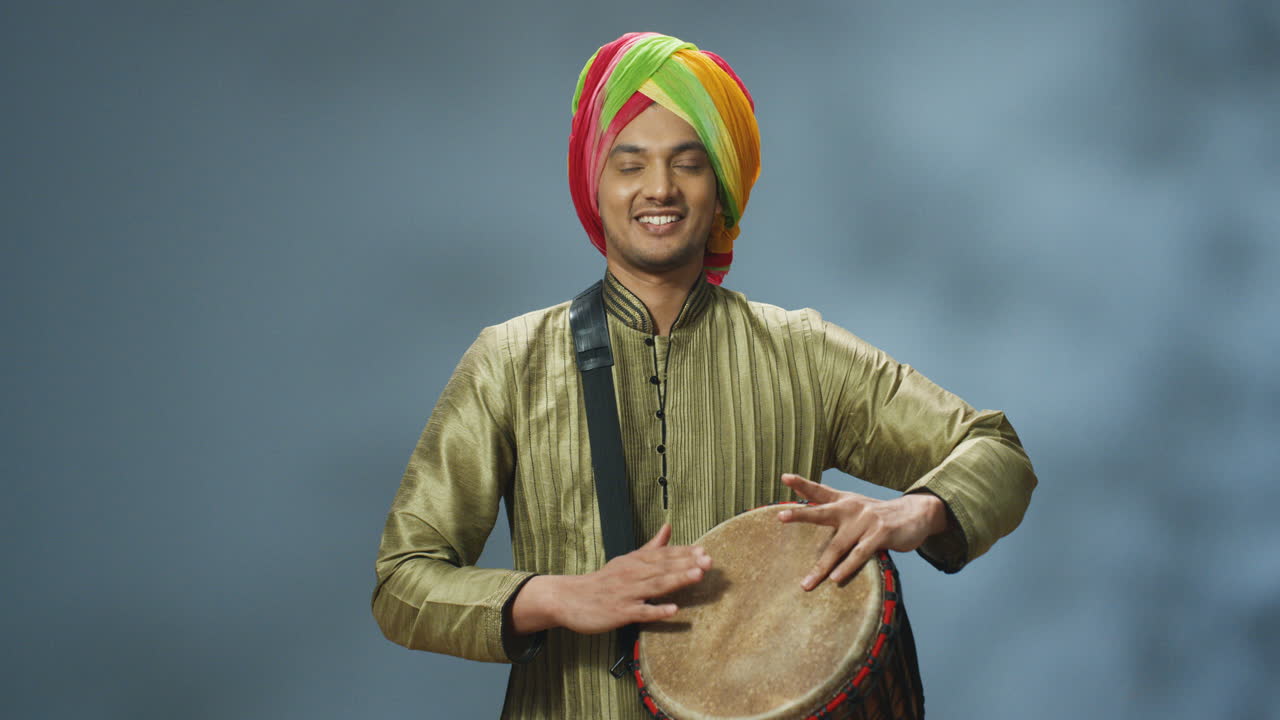 Portrait of young cheerful Indian man in traditional clothes and turban playing a drummer and smiling at camera
