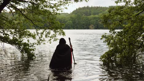 back view of grim reaper in black hooded robe walking into the water with scythe