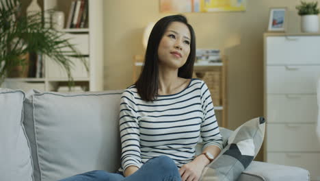 줄무늬 블라우스를 입고 거실에 앉아 소파에 앉아 카메라를 향해 행복하게 웃는 매력적인 젊은 여성