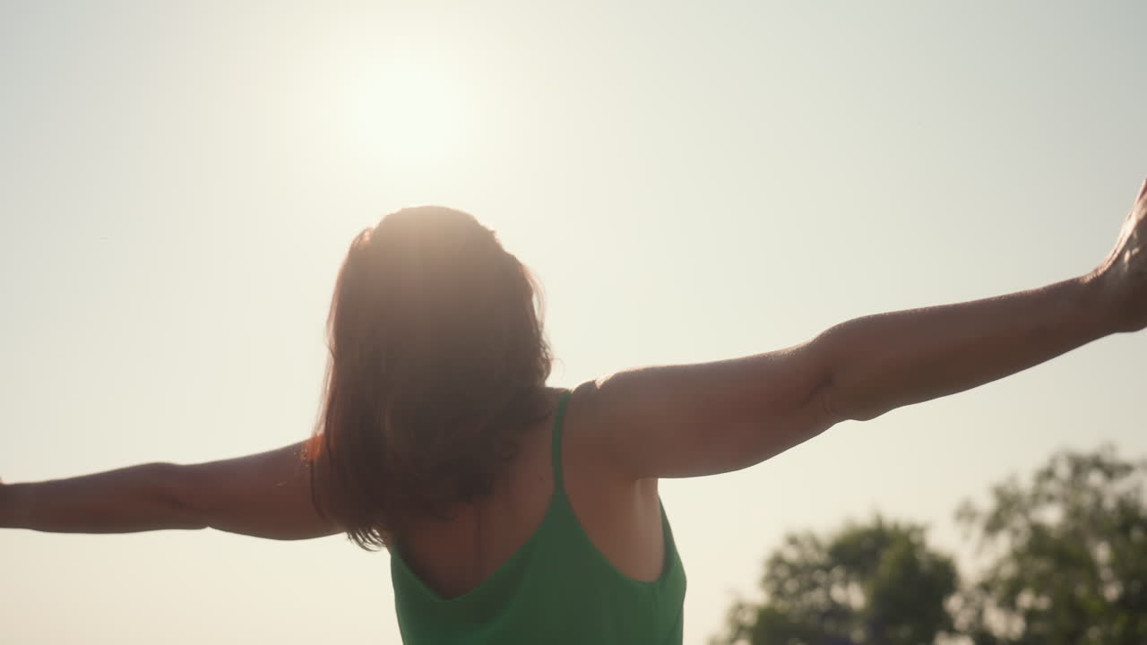 side view of woman in green dress lifting hand scratching hair under bright sky, face tilted upward with eyes closed, bathed in warm sunlight during peaceful outdoor moment with natural background