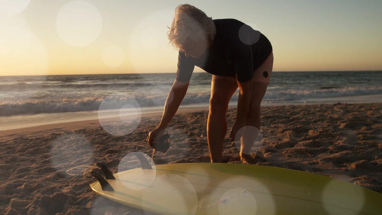 Seniorin beugt sich über ein gelbes Board am Ufer und passt die Surf-Leash an, mit Lichtreflex über den Händen.