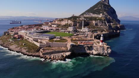 Waves-crash-against-the-rocks-below-the-red-white-colored-lighthouse-at-Europa-Point-in-Gibraltar-on-a-sunny-day-with-in-the-background-the-upper-rock-with-great-view