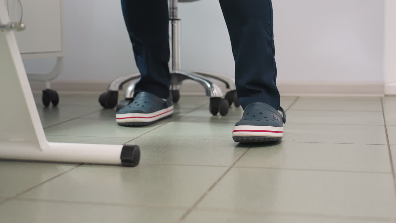 Leg view of beauty expert in Crocs with white socks wearing navy uniform standing from seat moving to side of massage chair in clean spa room with tiled floor, lamp and equipment trolley visible