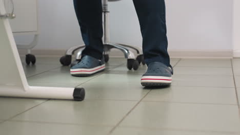 leg view of beauty expert in crocs with white socks wearing navy uniform standing from seat moving to side of massage chair in clean spa room with tiled floor, lamp and equipment trolley visible