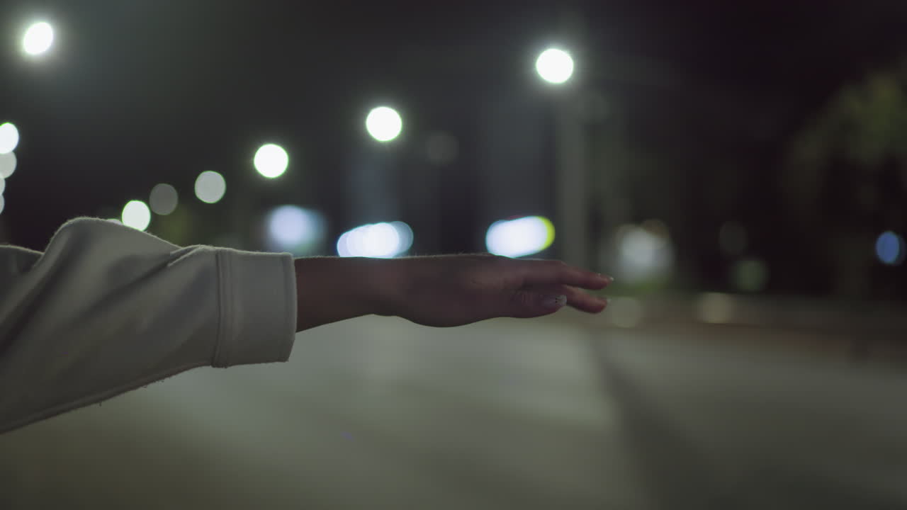 Close-up of outstretched hand wearing long sleeve coat reaching into quiet road under soft glow of streetlights during night in city, attempting to flag down taxi with vibrant bokeh background