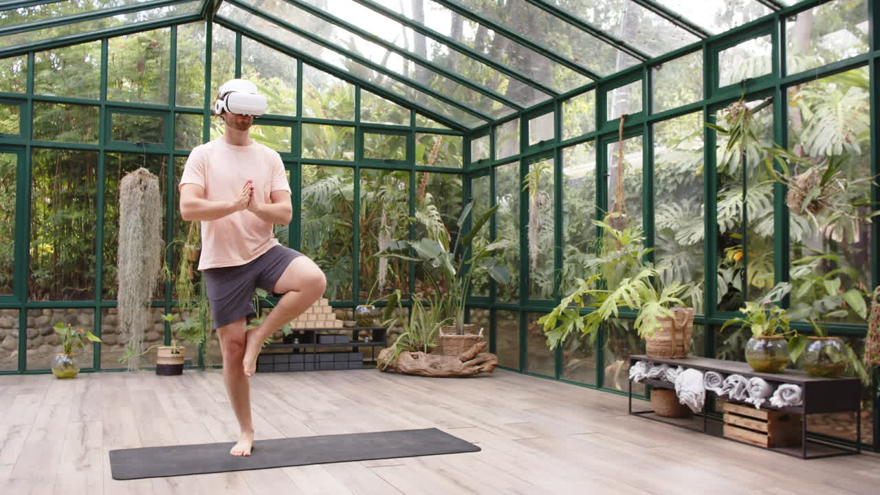 Practicing yoga, man wearing VR headset balancing on one leg in glass house, copy space