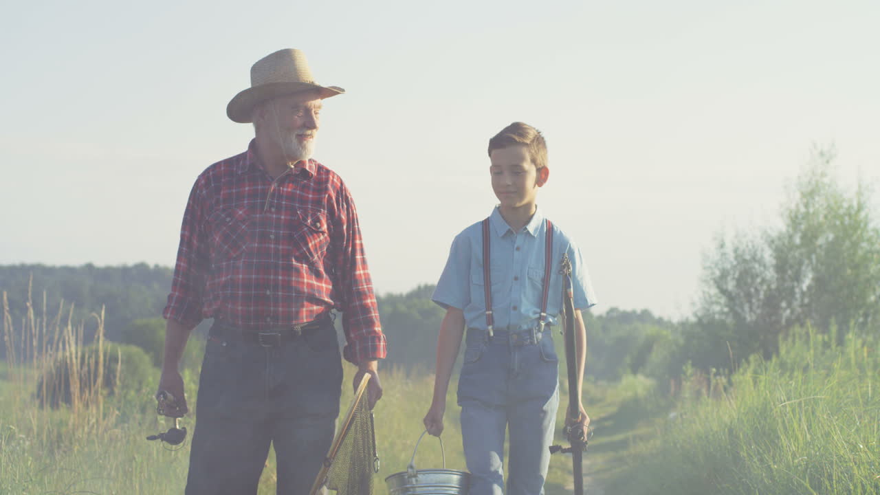 Caucasian grandfather and his little grandson walking together while holding fishing rods in the morning