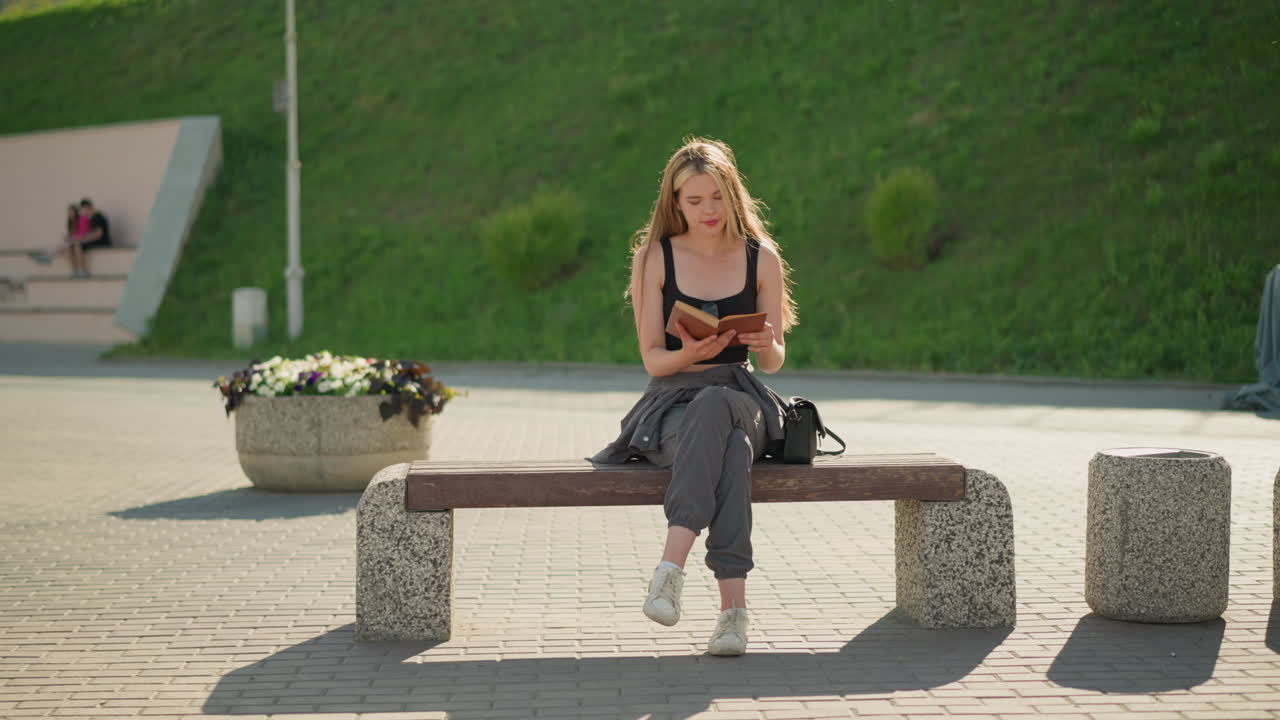 웃음으로 벤치에 앉아서 다리를 고 책을 읽는 여자, 그녀는 태양이 그녀를 비추는 동안 책을 부드럽게 들고, 배경에는 두 명의 멀리 떨어진 사람들이, 빛의 기둥이 있습니다.