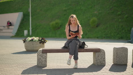 mujer sentada al aire libre en un banco con una sonrisa, leyendo un libro con las piernas cruzadas, sostiene el libro suavemente mientras el sol la ilumina, el fondo presenta dos personas distantes, un poste de luz