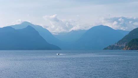 an aerial drone shot of a jetski sailing under the blue sky in lions bay, with serratus and tantalus mountain in the background