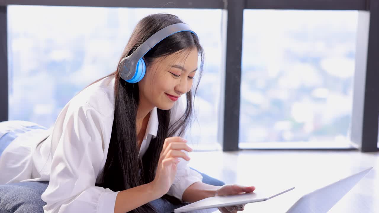 청춘과 귀여운 여성은 청바지와  셔츠를 입고 전자 장비 태블릿과 노트북과 함께 파란 헤드폰을 착용합니다. 여성은 아한 창 근처의 바닥에 누워 있습니다.