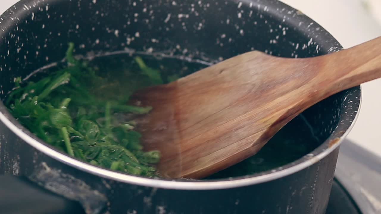 Using a wooden spatula to stir pot of boiling water blanching fiddlehead ferns for making pako salad, a local Filipino delicacy showing the authentic daily home life and culinary heritage