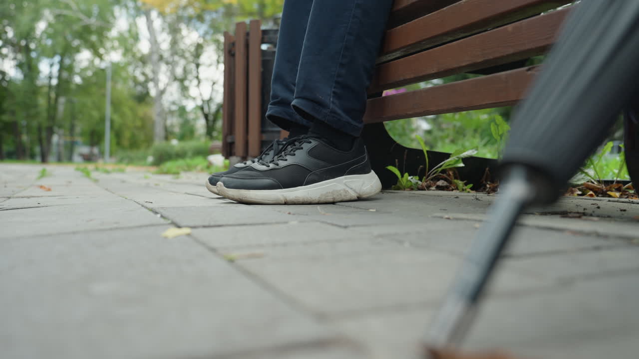 Nærbilde av parkbenk med joggesko og stokk, stemningsfull høstettermiddag med spredte blader, mann som sitter på benken og hviler foten, synlig jeansmansjett, uskarp stokkspiss i forgrunnen, liten dybdeskarphet