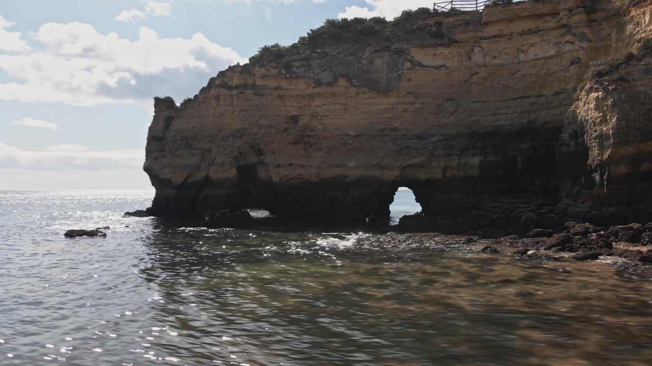Aerial drone performs a helix orbit around dramatic limestone sea arches and layered cliffs, with water sparkling in the sun near Lagos, Algarve, Portugal