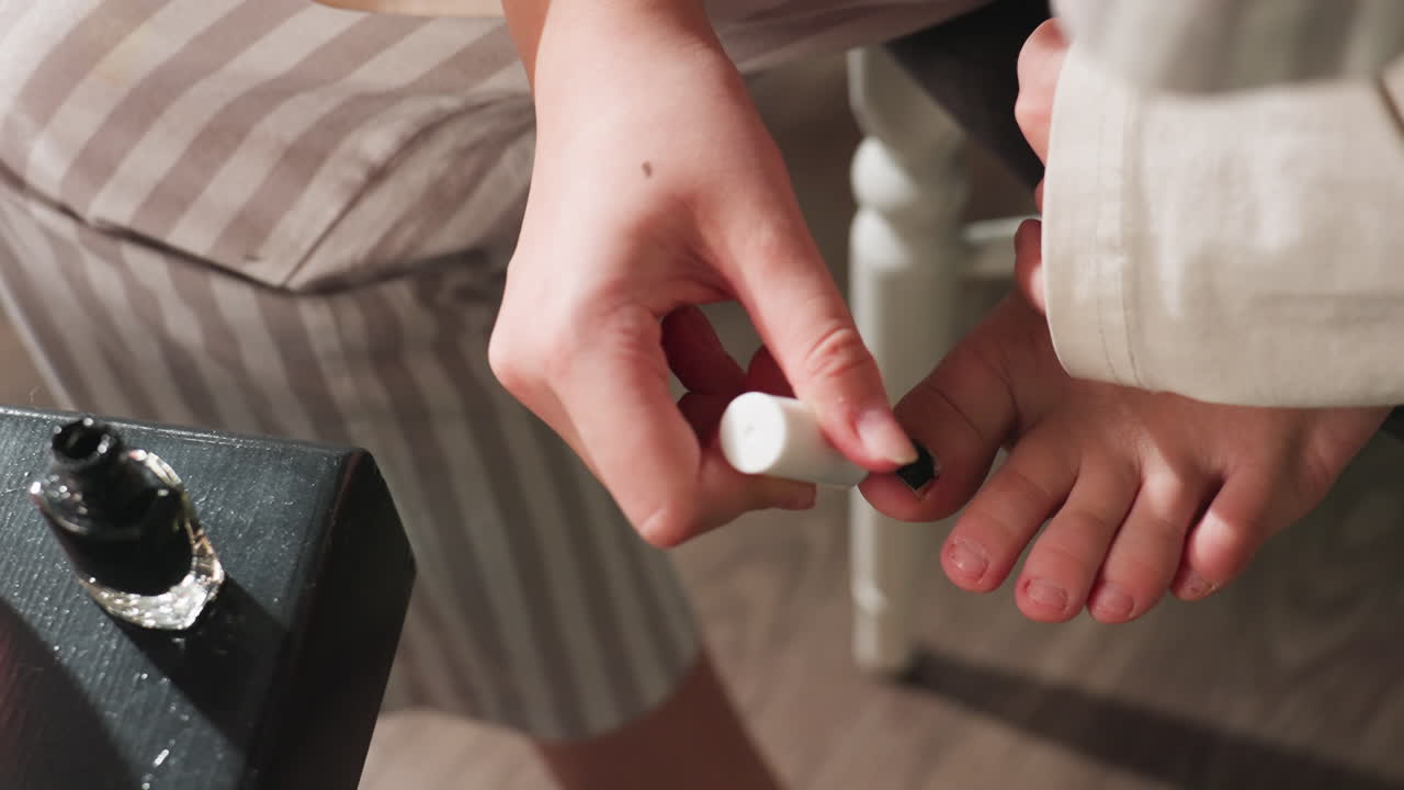 High angle view shows youth applying black polish to big toe nail with steady brush strokes, foot supported on chair, open bottle on table, home pedicure moment, focus on hygiene and beauty