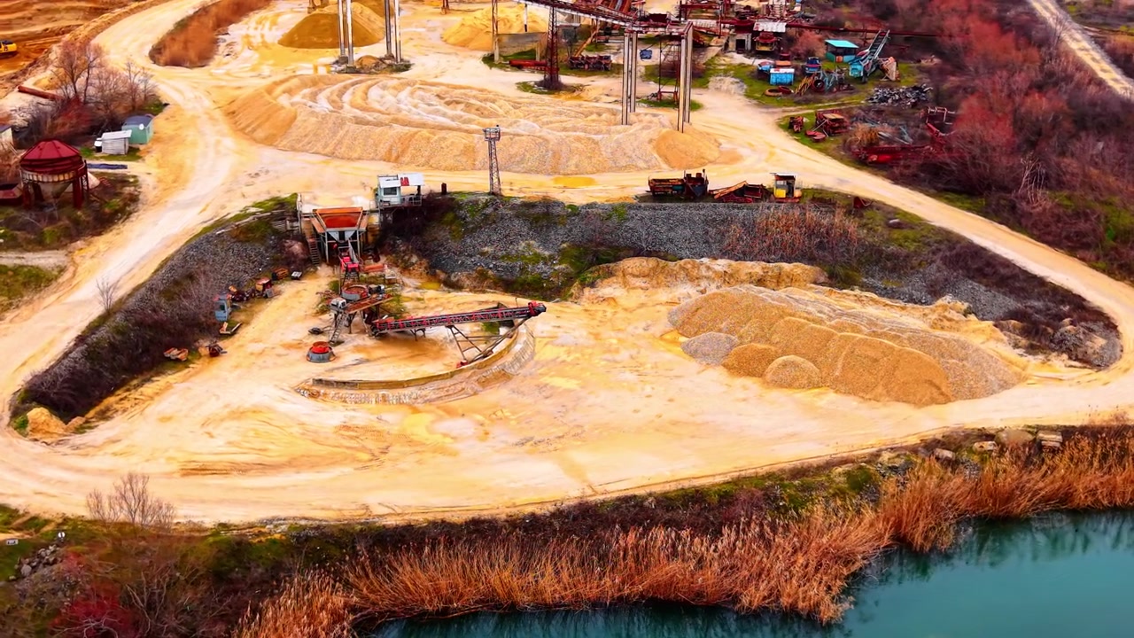 Elevated view of gravel processing facility beside quarry lake. Oblique drone shot showing conveyor, gravel stockpiles, circular sand mound, reed waterfront and teal lake at golden hour.