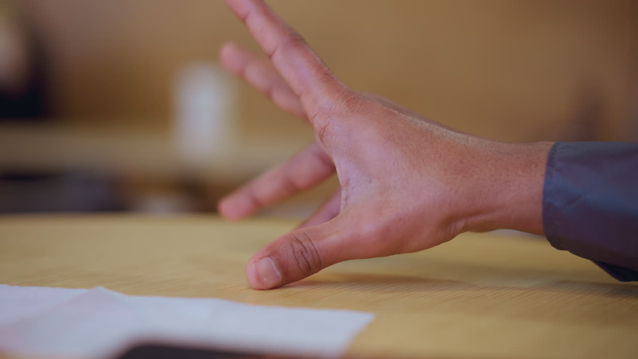 Close-up of young man's hand gently placed on wooden table near white napkin, fingers slightly bent, suggesting moment of contemplation, tension, or hesitation in warm indoor environment