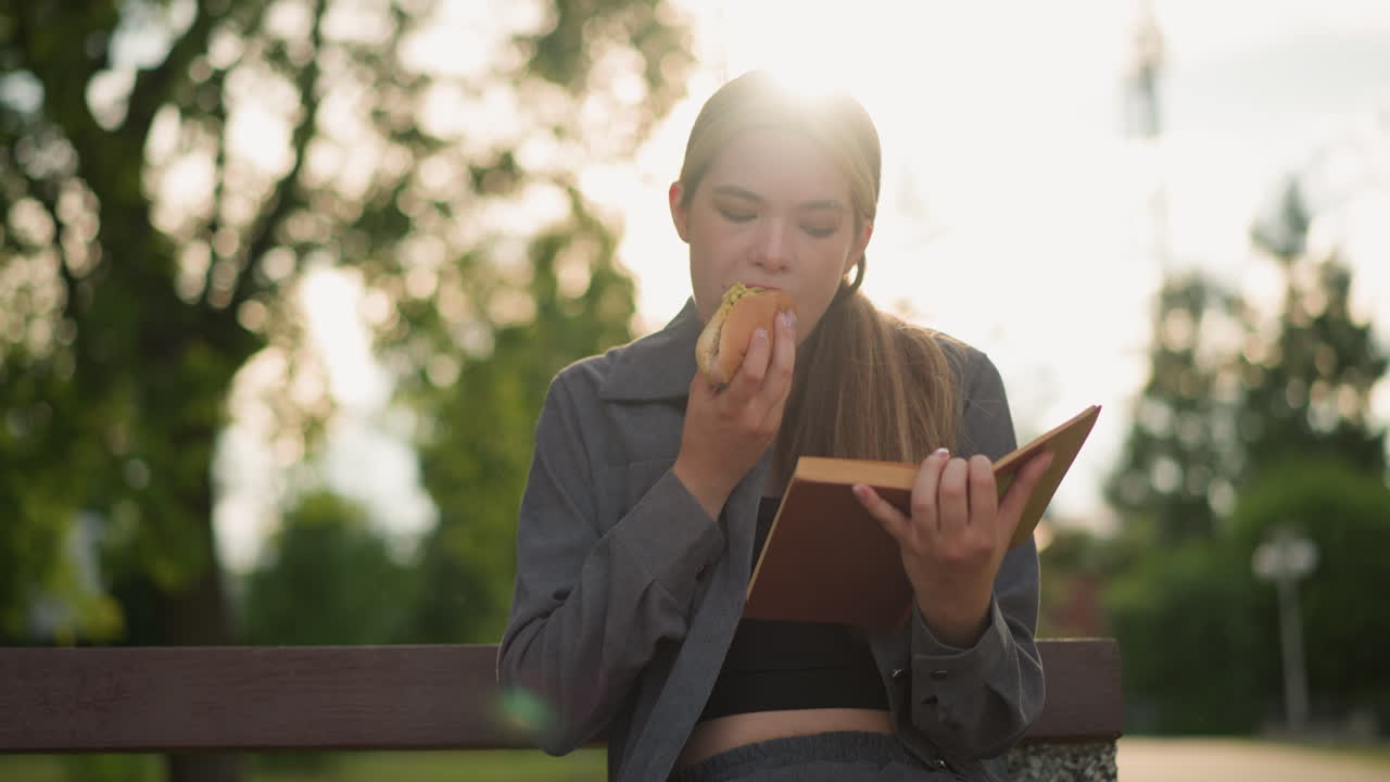 회색 을 입은 여자가 야외에서 책을 읽는 동안 간식을 먹고, 나무와 초록색으로 둘러싸인 공원 벤치에 앉아, 흐릿한 배경으로 부드러운 빛이 비추고 있습니다.