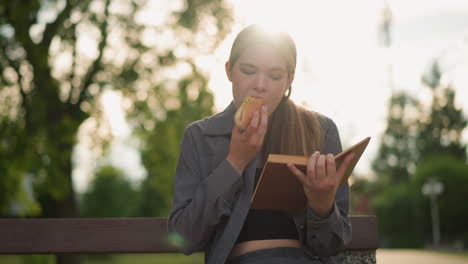 dama de camiseta gris comiendo bocadillo mientras lee un libro al aire libre, sentada en un banco del parque rodeada de árboles y vegetación con fondo borroso, la suave luz del sol ilumina