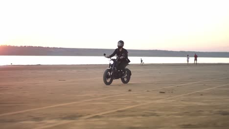 Biker-in-helmet-and-jacket,-man-is-riding-at-sport-motorbike.-Wind-blowing,-water-front,-sunset