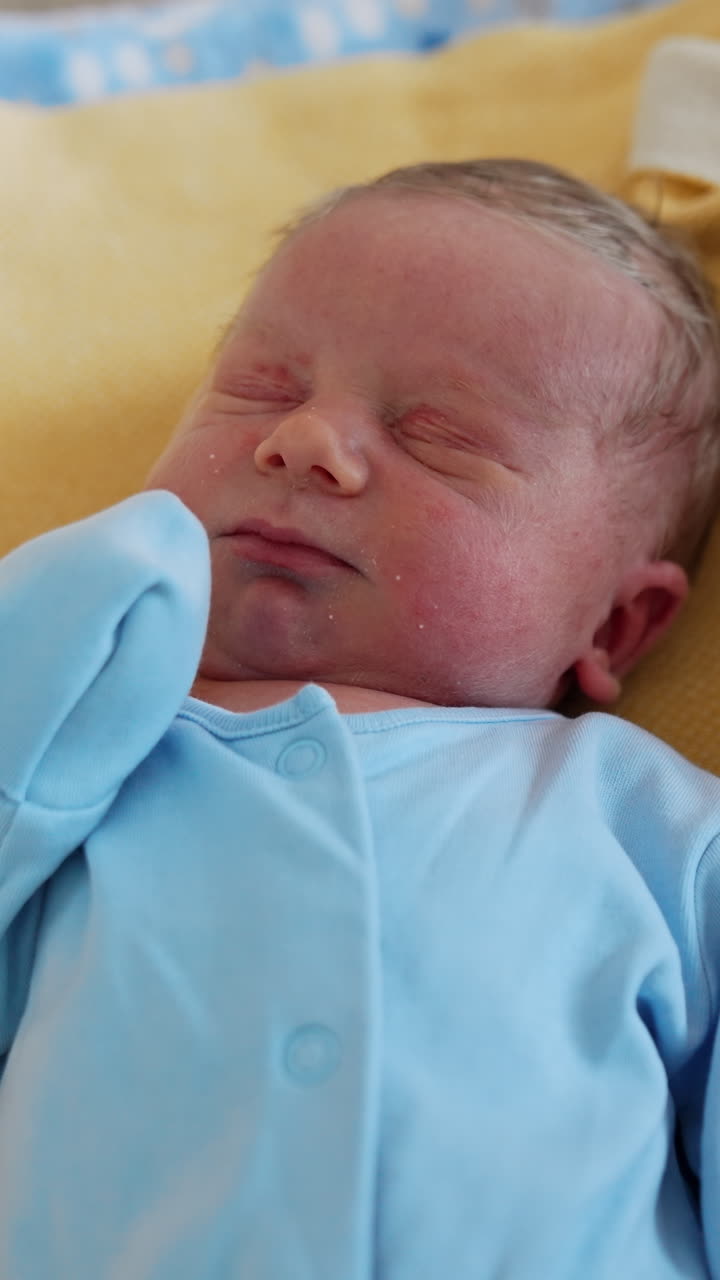 Adorable newborn boy with fair hair. Little baby in a blue bodysuit sleeping after birth in hospital. Close up. Vertical video