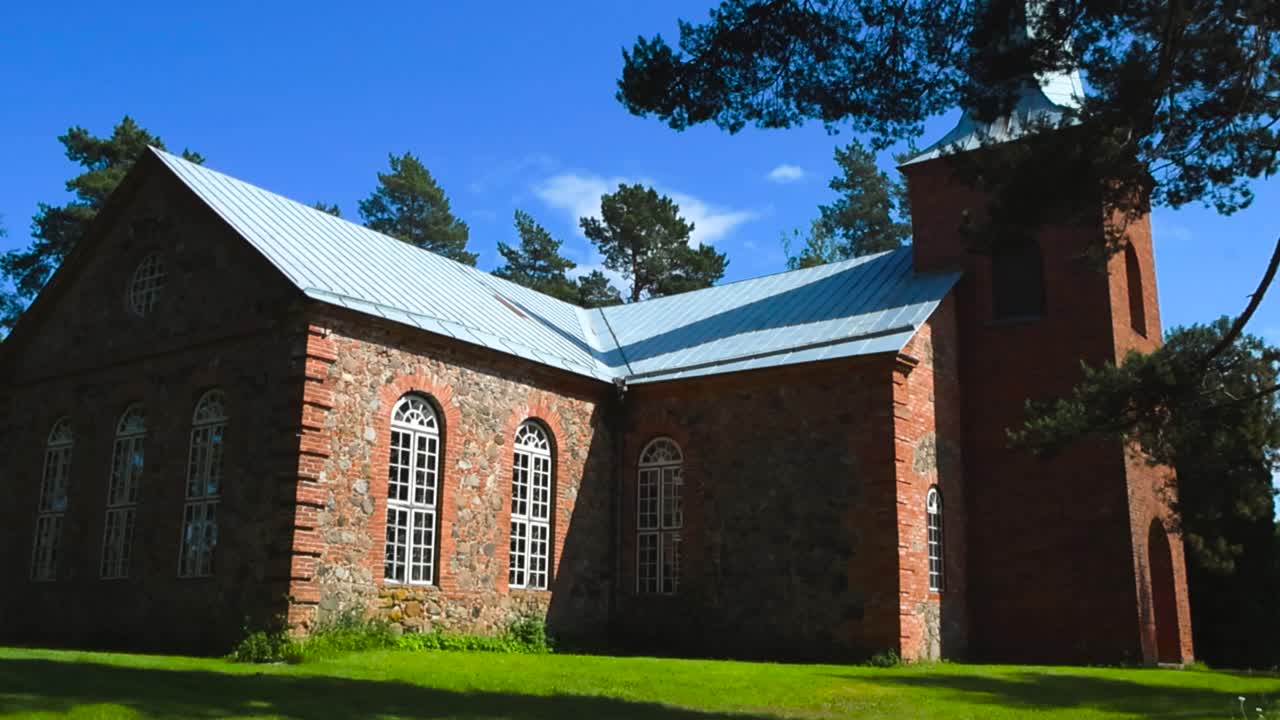 Gorgeous low angle footage of a red colored old and vintage historic brick chursh in the middle of the woods or a forest during a summer sunny day with blue sky, white clouds and treetops in the back