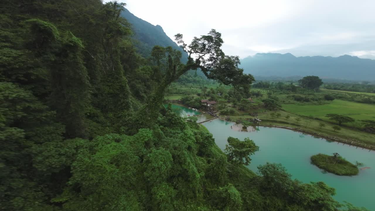 Drone descends tree tops above Blue Lagoon, Vang Vieng, Laos, with green landscape and limestone hills under overcast skies
